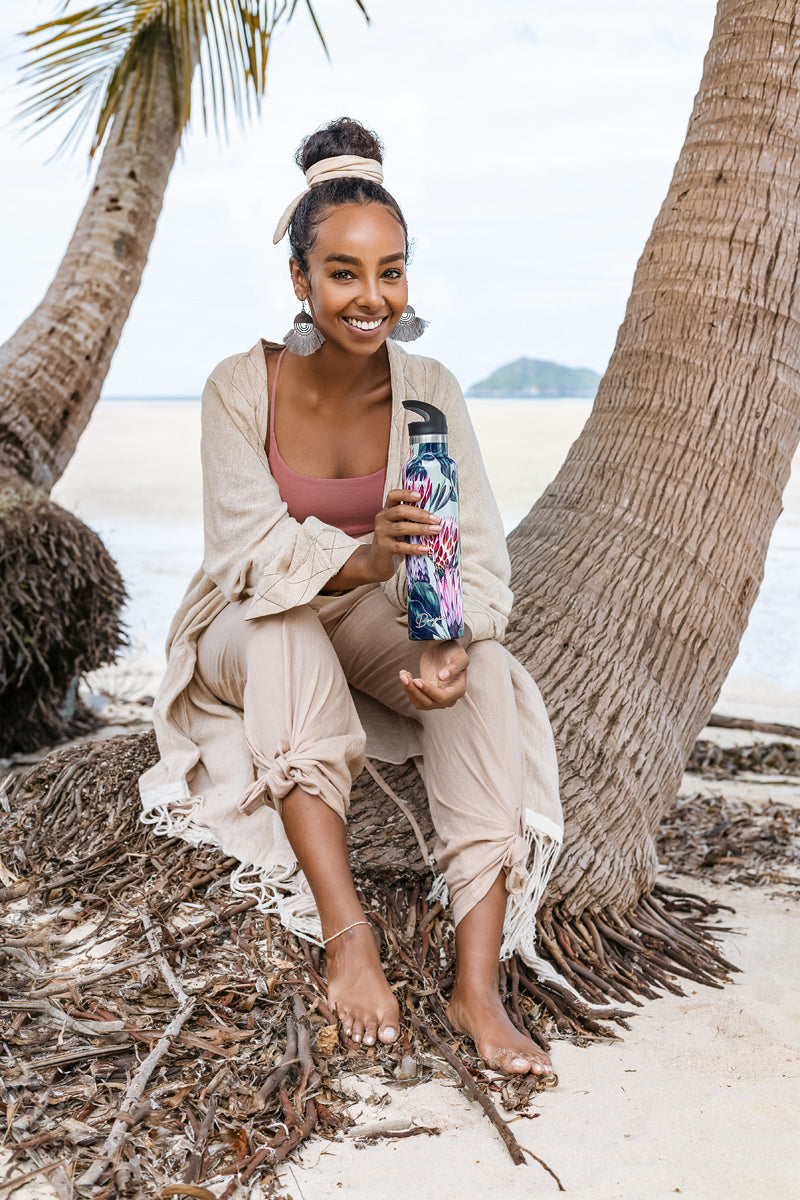 elegant African model holding protea water bottle smiling on a beach sitting under palm tree on a beach. 